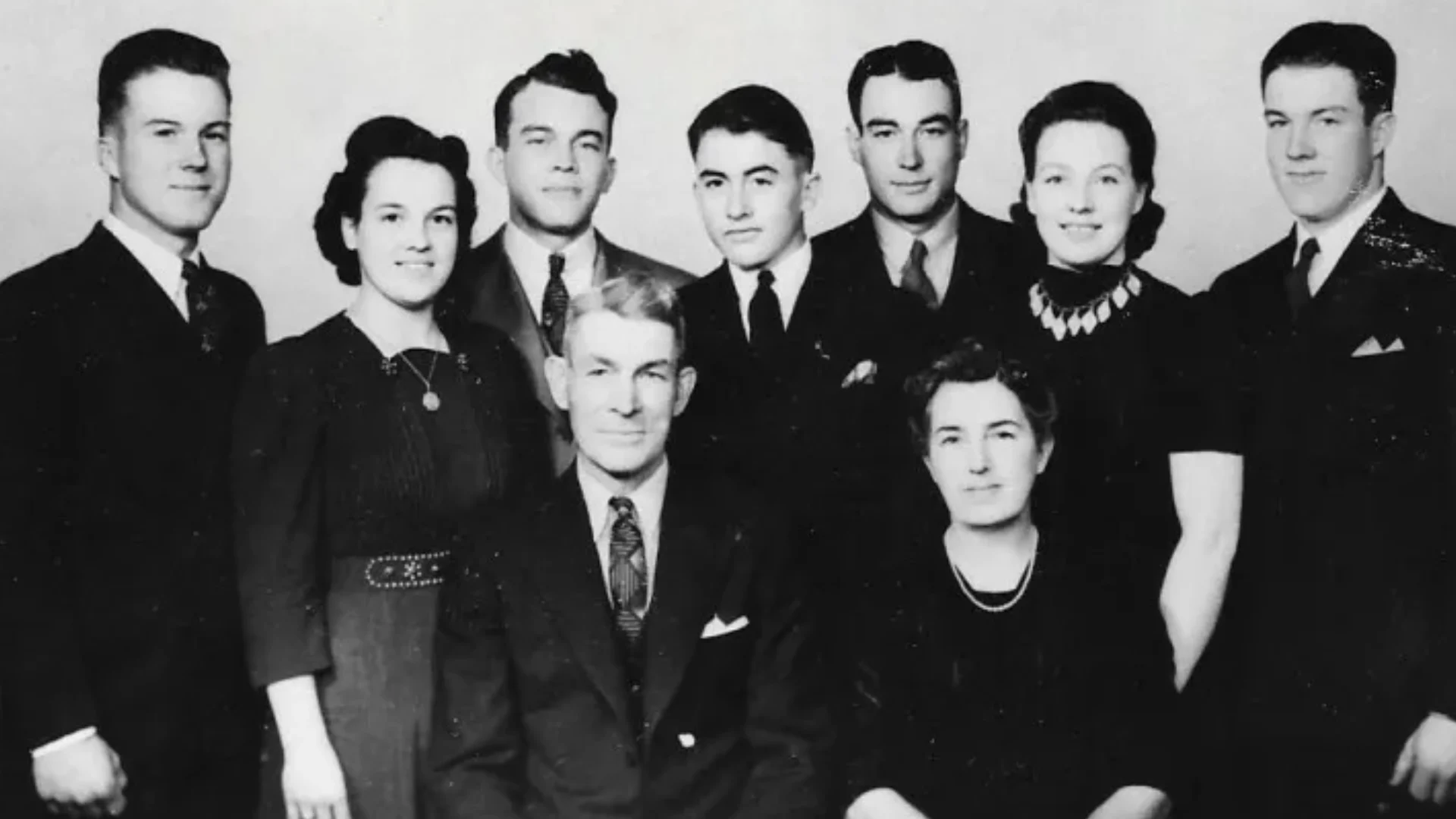 Black-and-white formal group portrait of nine relatives in suits and dresses, posed in two rows in a studio setting.