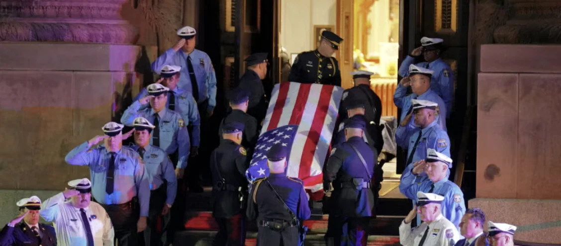 Casket draped in the American flag is carried out by uniformed police and military personnel while they salute at a ceremonial funeral entrance.