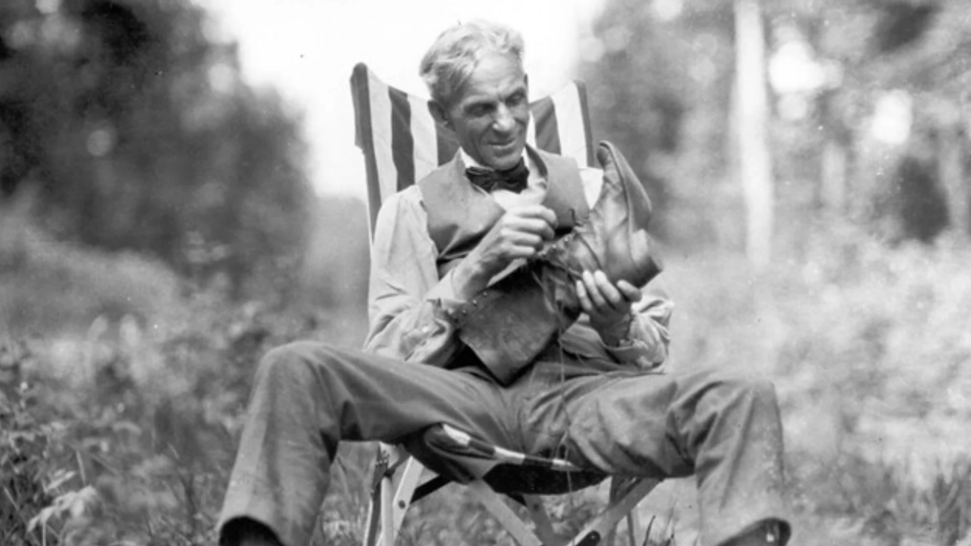 Man in a vintage suit sits in a striped outdoor chair, inspecting a boot in his hands outdoors.