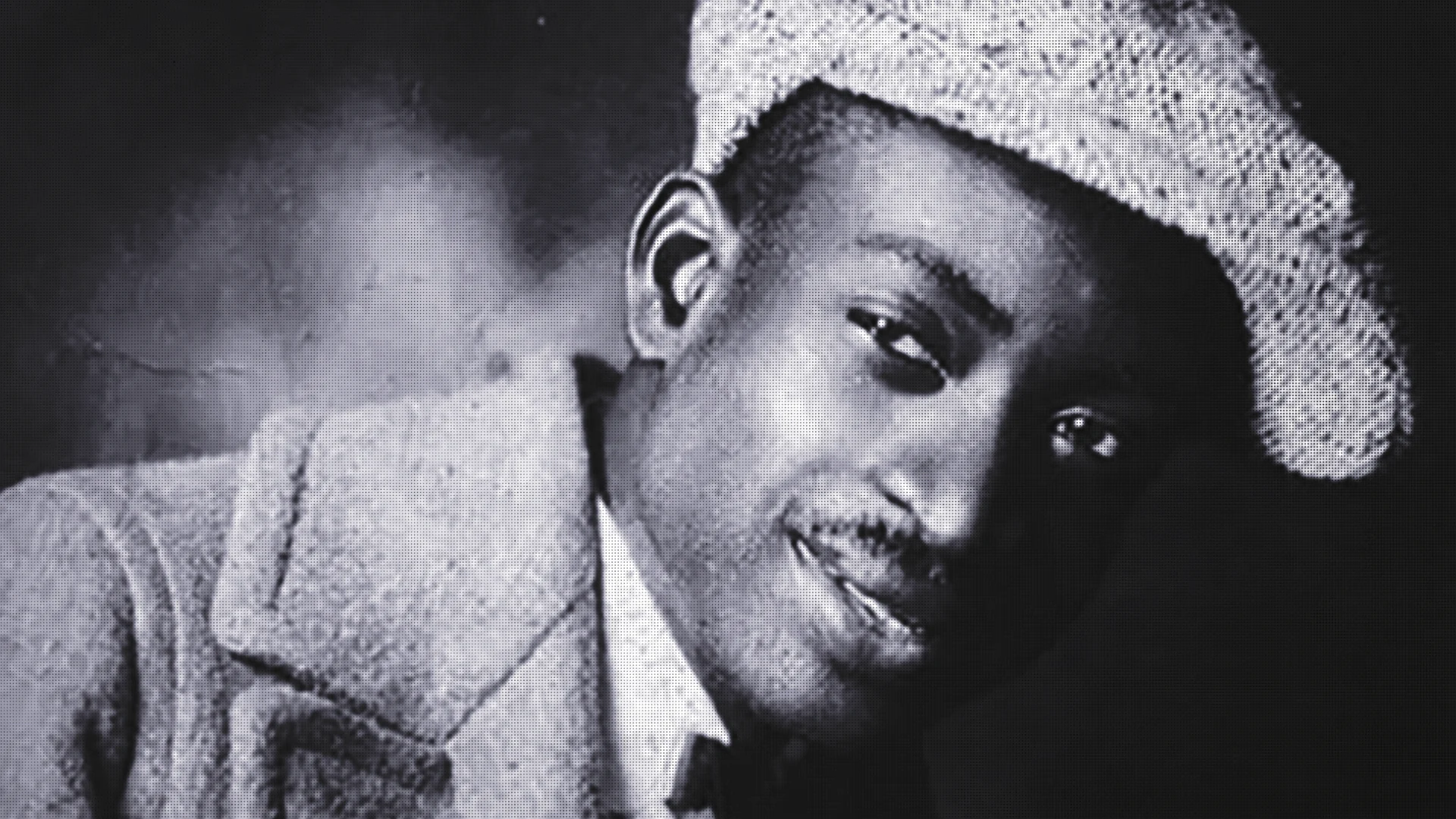 Black-and-white close-up portrait of a smiling man wearing a textured hat, looking toward the camera.