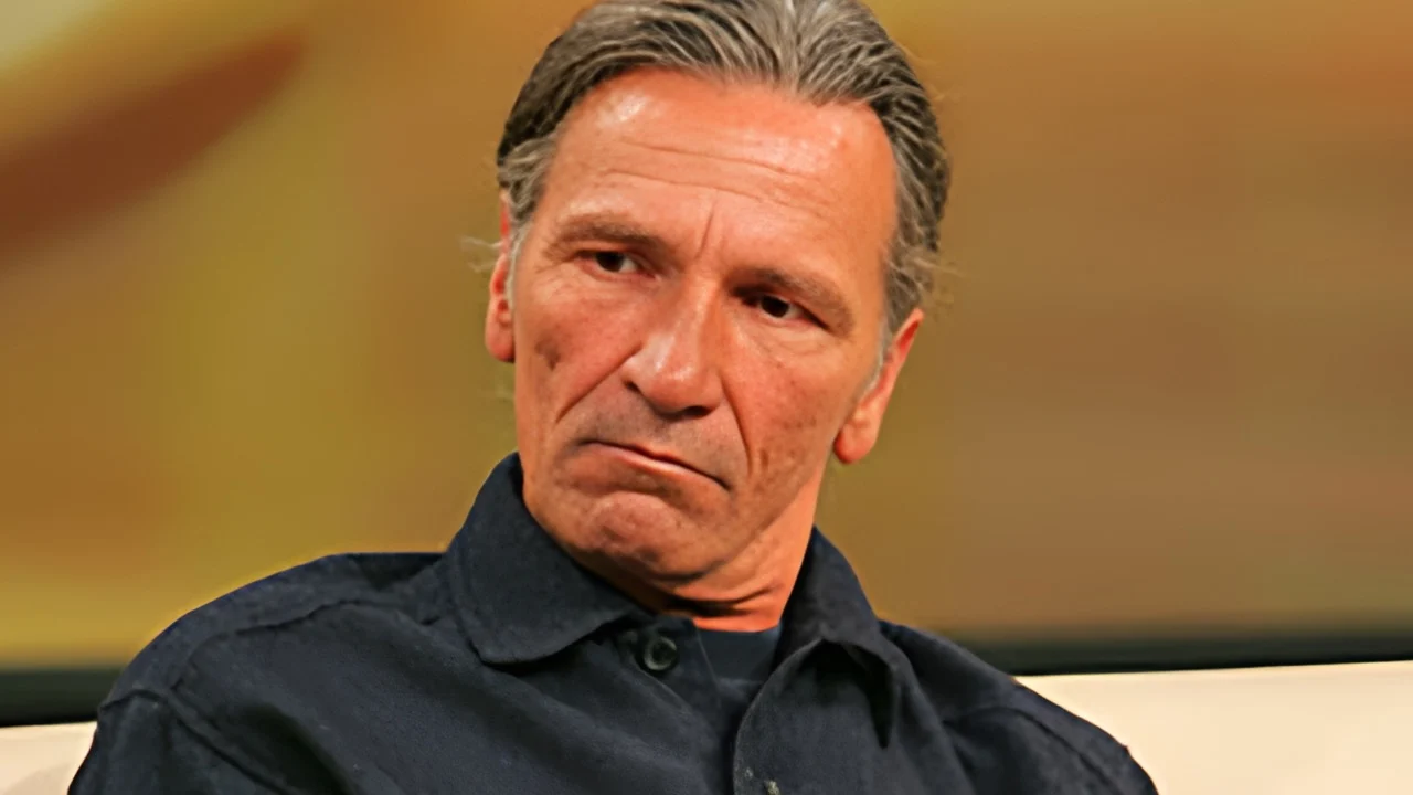 Middle-aged man with gray hair wearing a dark shirt, giving a thoughtful, somber look.