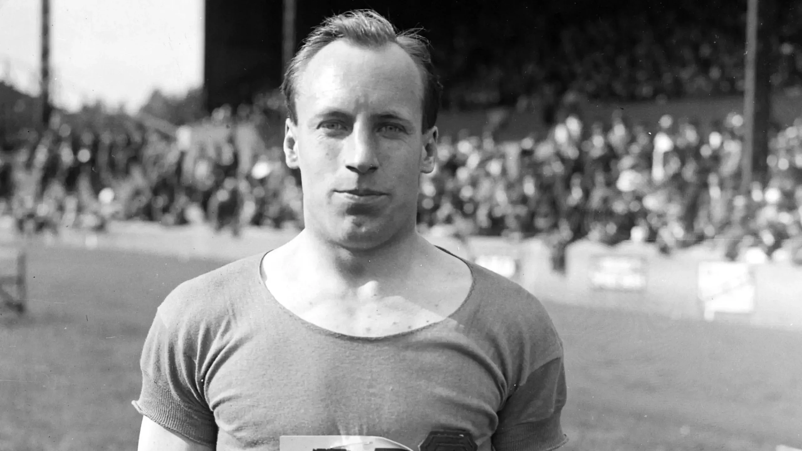 Man wearing a short-sleeve shirt stands outdoors, with a blurred crowd in the background.