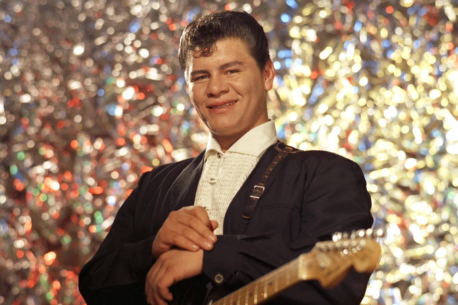 Smiling man in a white shirt and dark jacket holding a guitar on a glittery stage backdrop with colorful lights.