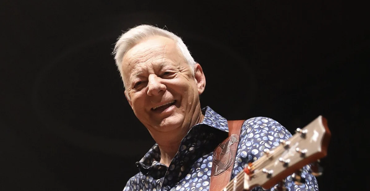 Older man with white hair smiling while playing an acoustic guitar on stage.