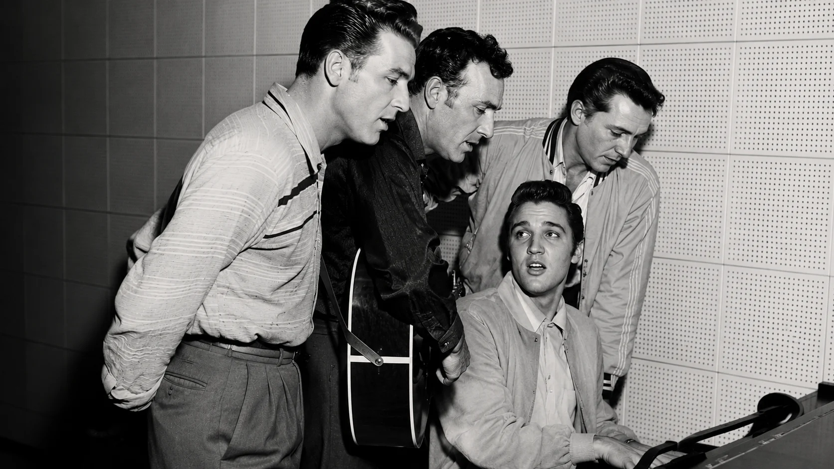 Four men in a recording studio gather around a piano; one sits at the keyboard, another holds a guitar, taken in black and white.