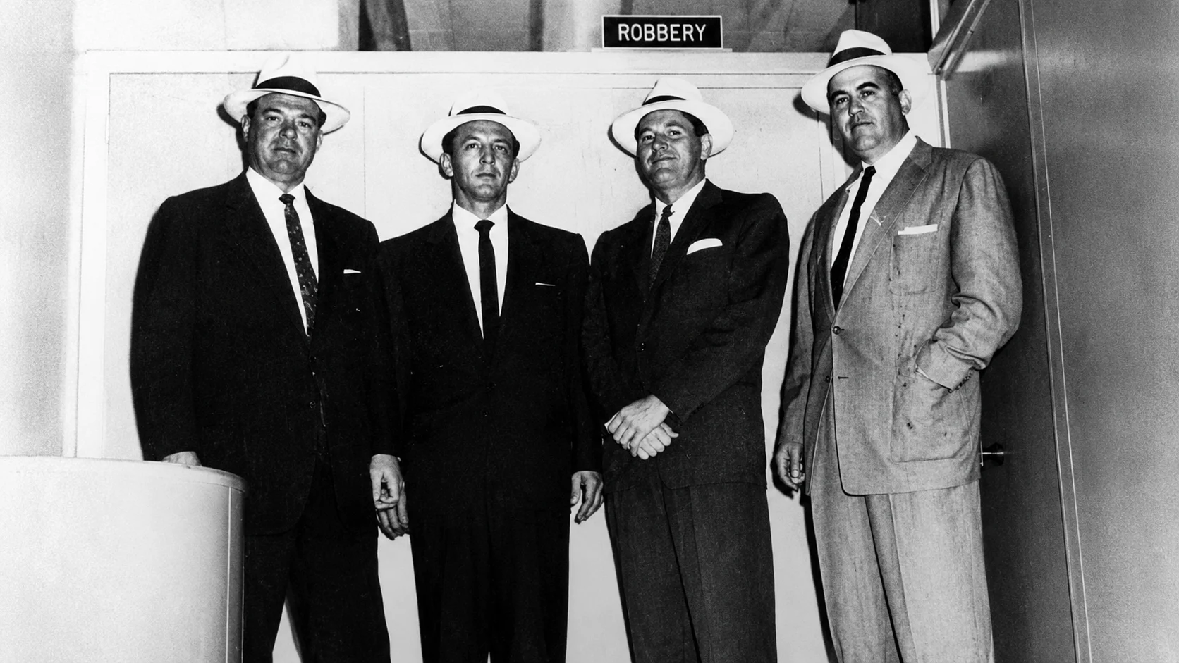 Four men in 1940s-style suits and fedoras stand together in a hallway beneath a sign that reads 'ROBBERY'.
