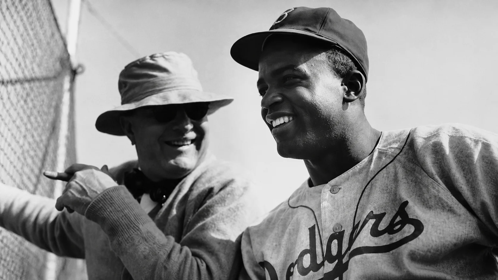 Two baseball players in vintage Dodgers uniforms smile and chat beside a chain-link fence.