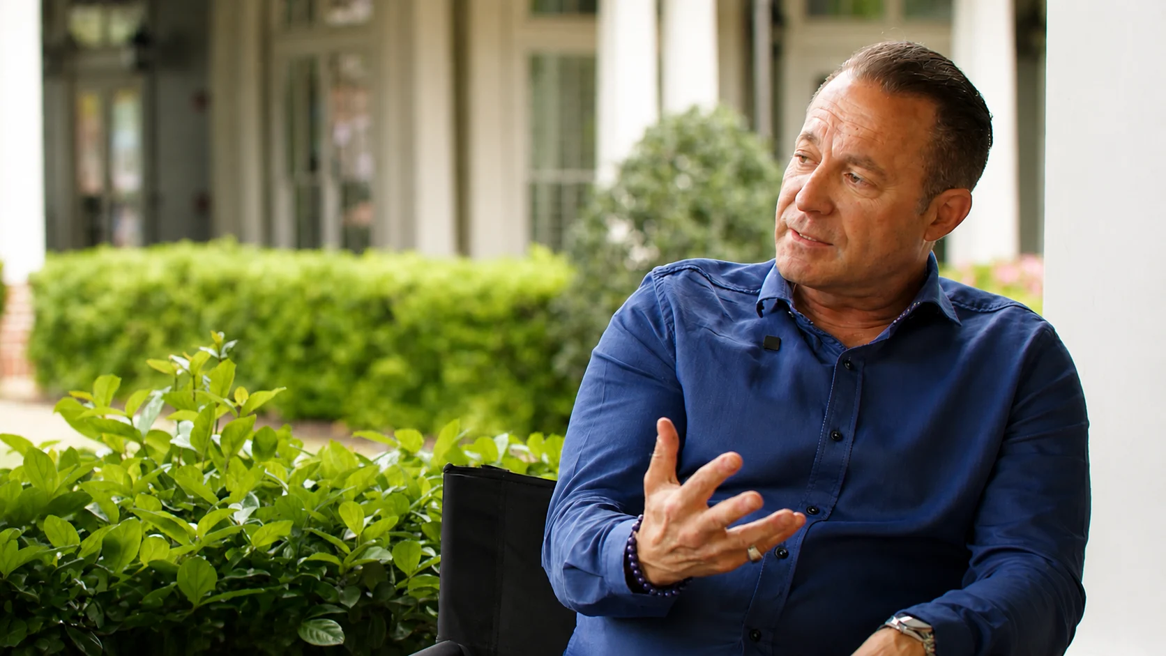 Man in a blue button-down shirt sits outdoors, gesturing with his hands while talking, with green bushes in the background.