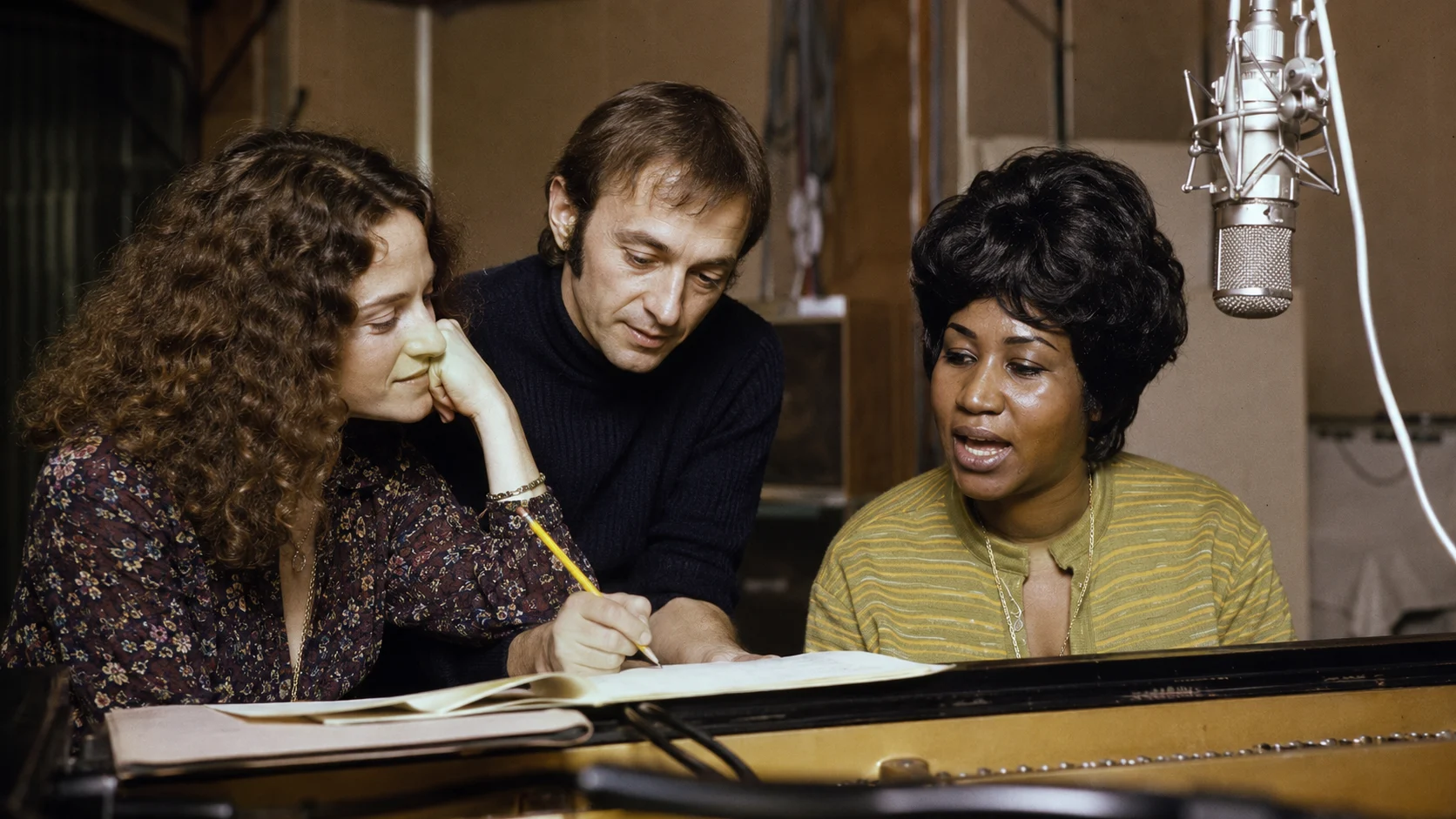 Three musicians gathered around a piano, looking at sheet music as a large studio microphone hangs above them in a recording set‑up.