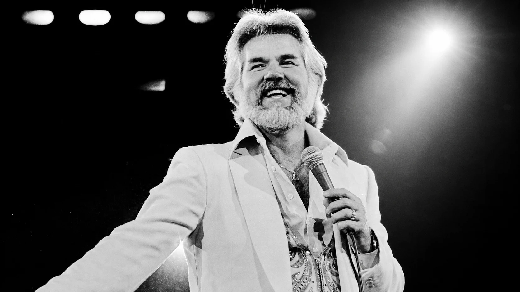 Smiling male singer with white hair and beard, in a white suit, holding a microphone on stage under bright lights.