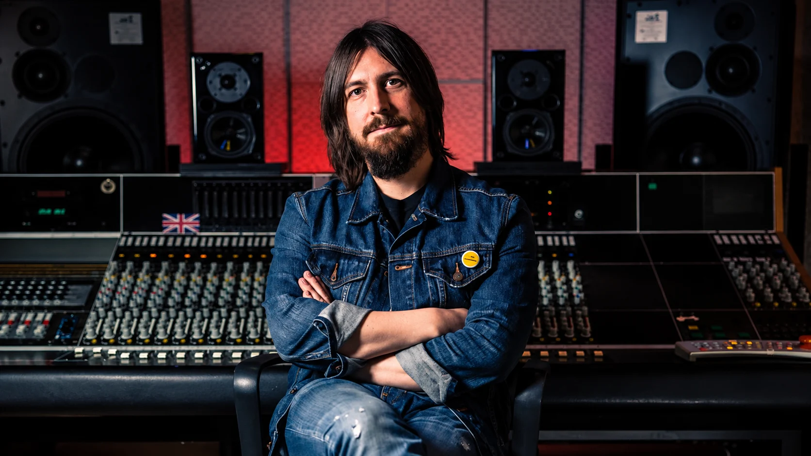 Portrait of a man with shoulder-length hair and beard seated with arms crossed in a recording studio, surrounded by a large mixing console and studio monitors in a dimly lit room.